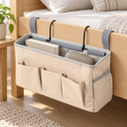 Beige bedside caddy organizer hanging from a light wood bed frame with books and daily essentials